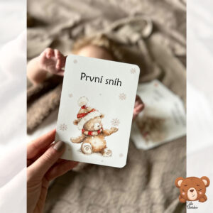 Maminka drží vánoční milníkovou kartičku První sníh s medvídkem v čepici vedle miminka ležícího na dece – součást autorské kolekce Moje první Vánoce od Julie Vanhaber.